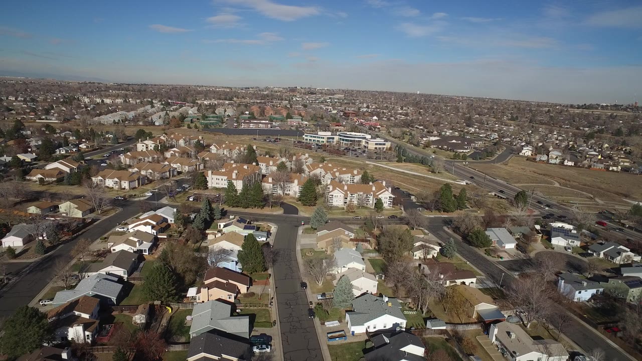 Opening aerial frame over Hidden Lake, Colorado, with foreground homes, a curving residential street, open brown land, distant buildings, and no visible people.
