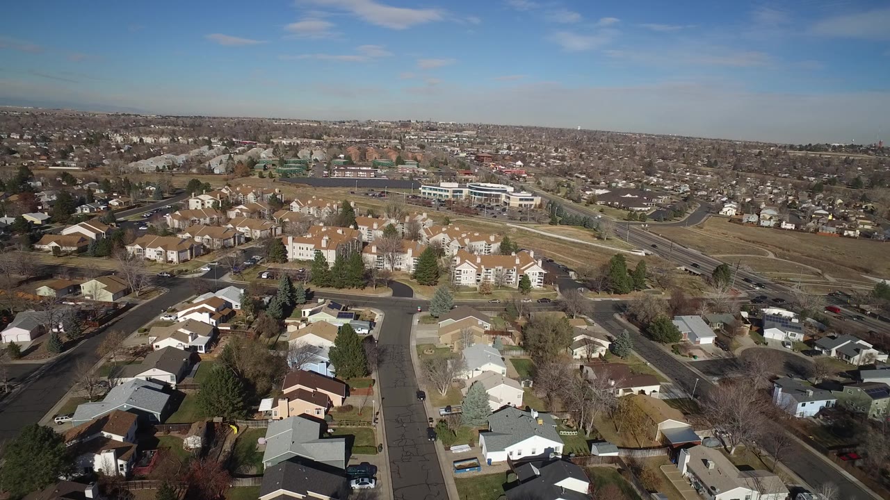 Second aerial frame in Hidden Lake, Colorado, showing clustered rooftops below, a broad road at right, brown open ground, distant housing, and no visible people.