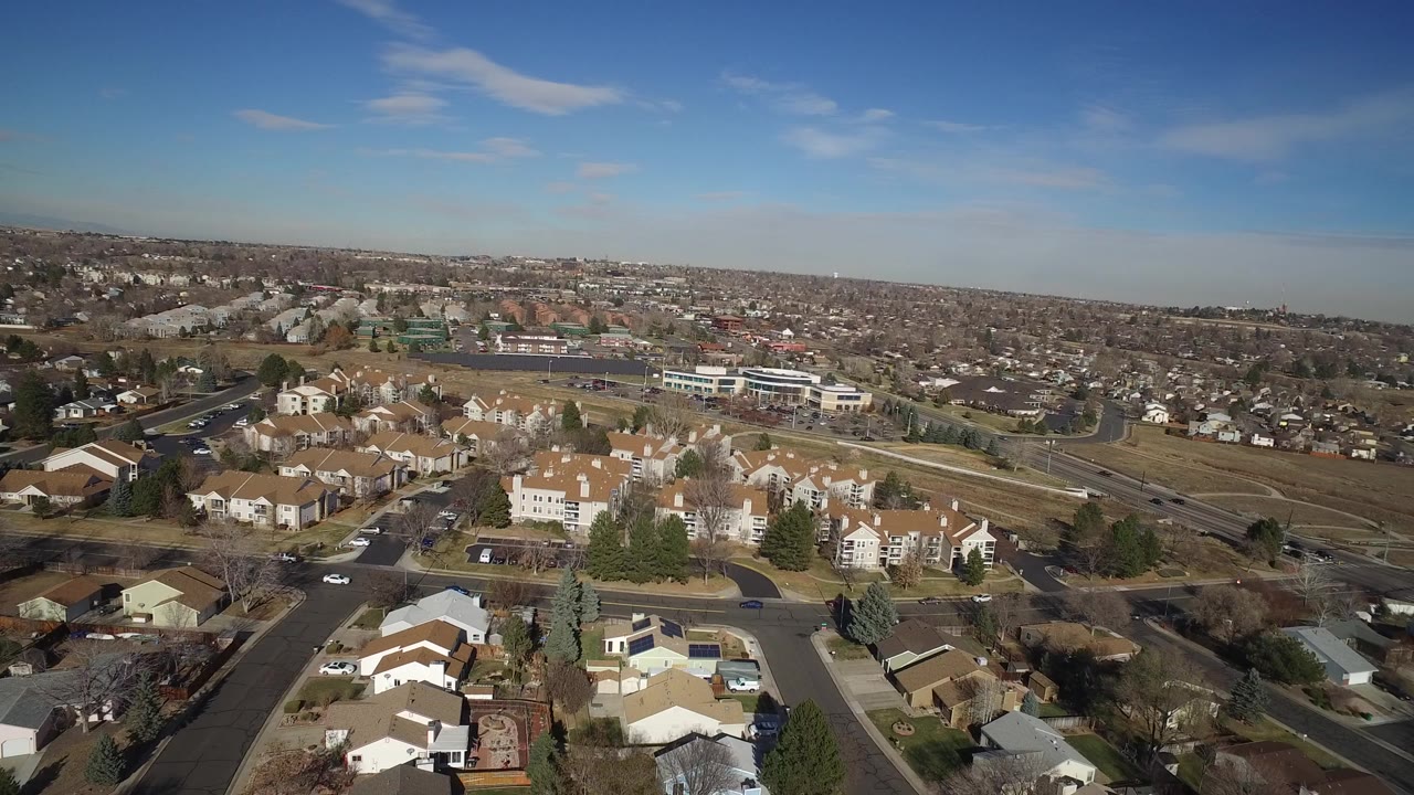 Aerial view over suburban homes, streets, trees, and distant rooftops.