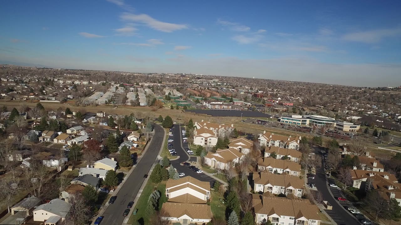 A high aerial view in Hidden Lake, Colorado looks over tan-roofed homes, curving streets, leafless trees, distant housing, and a blue sky with no visible people.