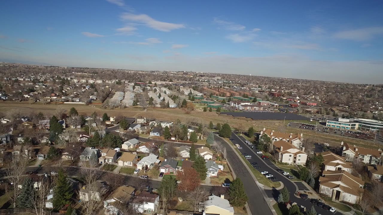 Opening aerial frame over Hidden Lake, Colorado, with houses and leafless trees in the foreground, a road with cars, open brown fields, distant neighborhoods, blue sky, and no visible people.