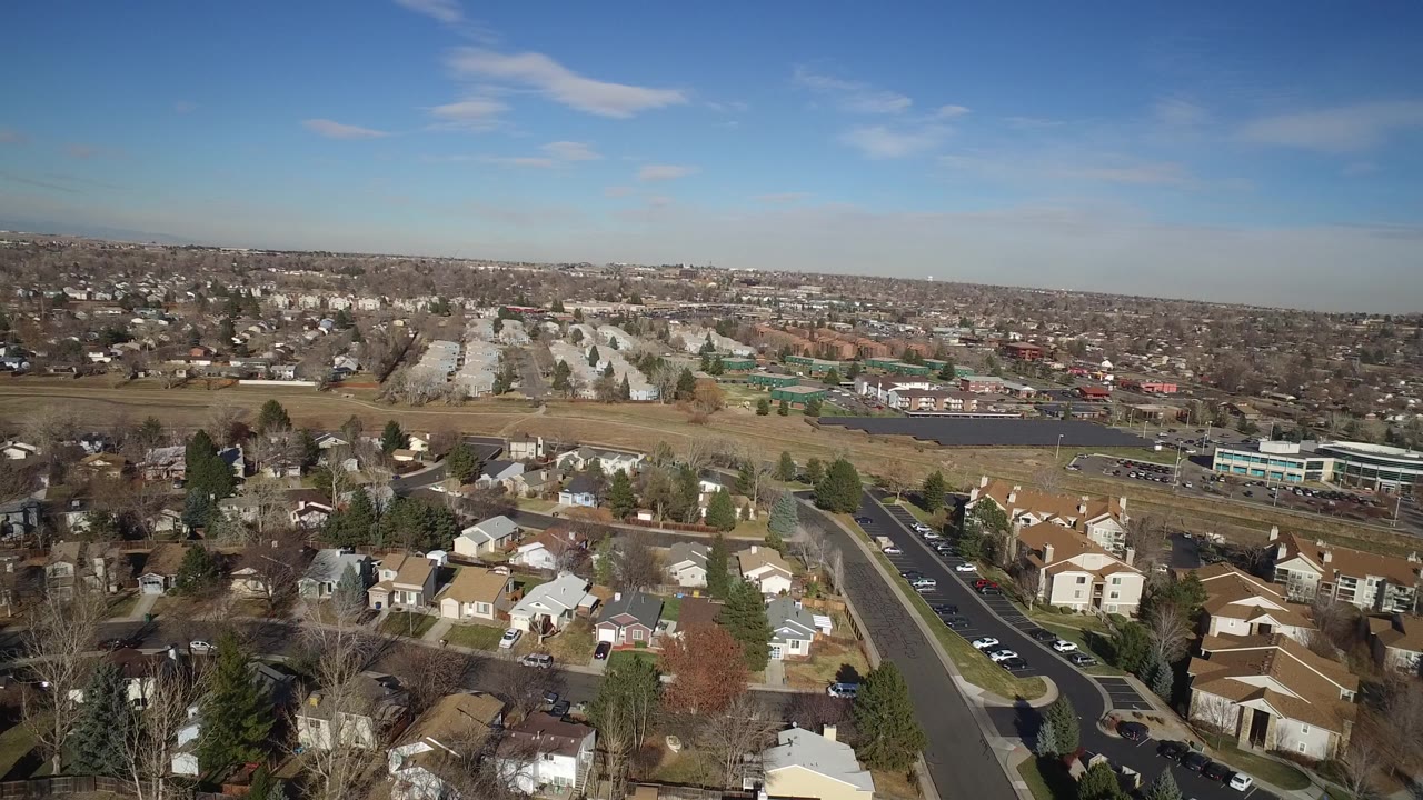 Hidden Lake, Colorado aerial at 0:01 showing foreground homes, a diagonal road, parked and moving cars, open fields, apartment rows in the midground, and no visible people.