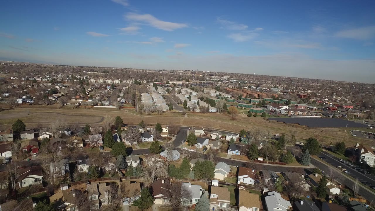 Opening aerial frame over Hidden Lake, Colorado, showing foreground homes, a broad grassy area, a small body of water, distant residential blocks, and no visible people.