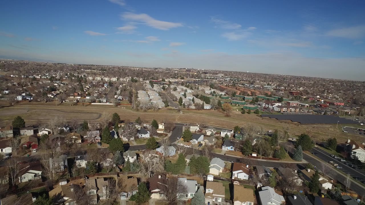 Aerial frame from Hidden Lake, Colorado, with more foreground rooftops visible below, open grass and a pond or small lake across the middle, and no visible people.