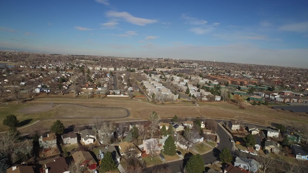Aerial view in Hidden Lake, Colorado with rooftops at the bottom edge, a curved road and field in the midground, distant housing blocks, and no visible people.