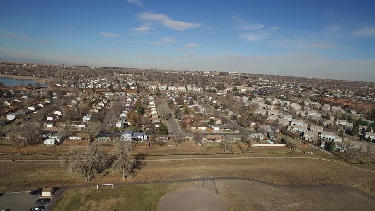 Aerial frame over Hidden Lake, Colorado showing an open field in the foreground, rows of small homes beyond it, blue lake water at left, and no visible people.
