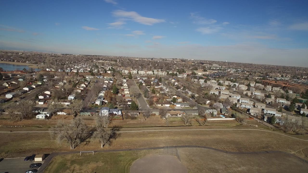 Aerial frame over Hidden Lake, Colorado with straight neighborhood roads centered in view, clustered houses, winter-brown lawns, lake water on the left, and no visible people.