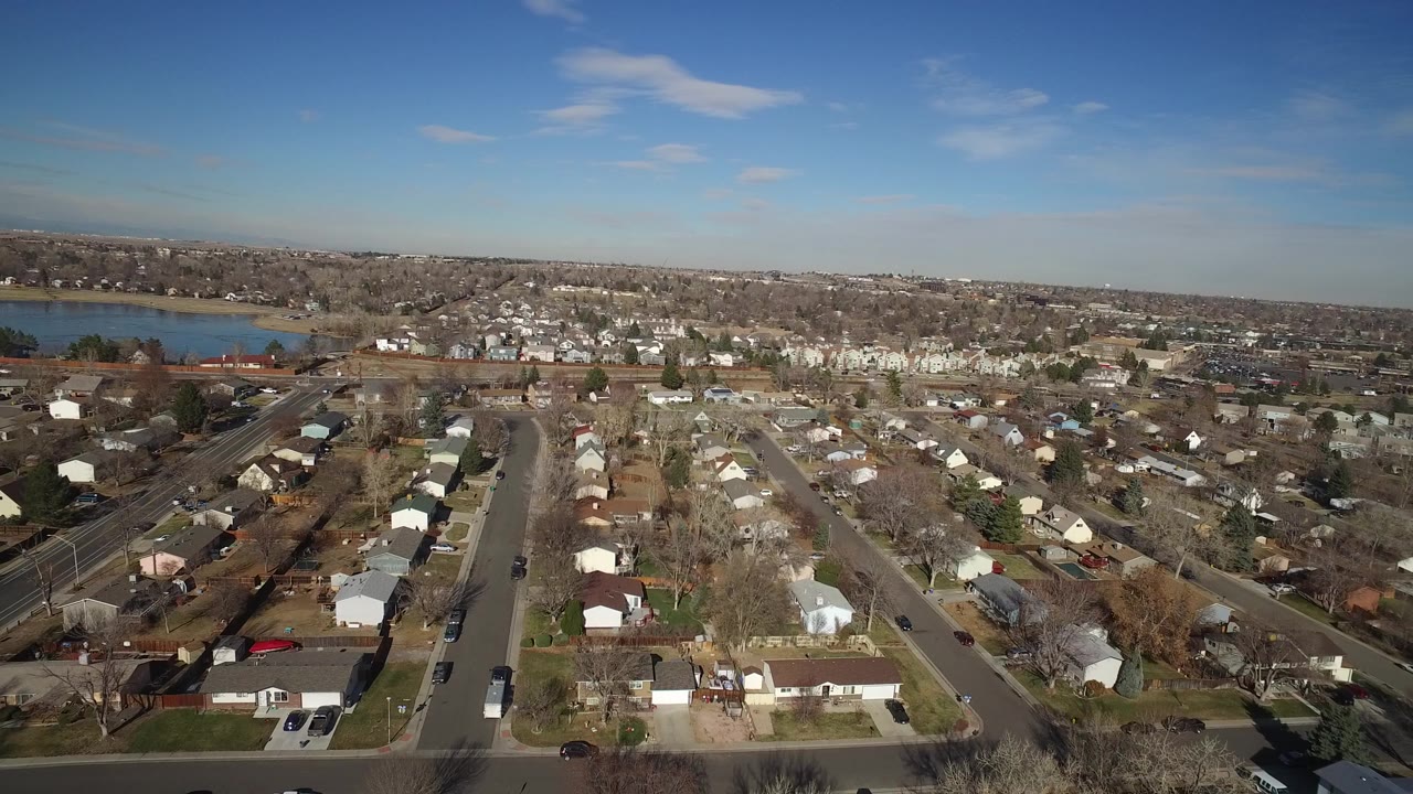 Opening aerial frame over Hidden Lake, Colorado, with residential streets and small houses in the foreground, a blue lake beyond the neighborhood, and no visible people.