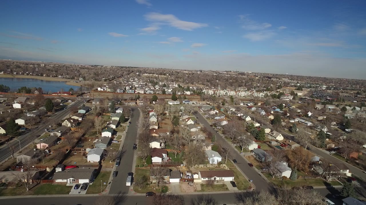 Aerial view over Hidden Lake, Colorado, showing a straight neighborhood street, rows of small homes, bare trees, the nearby lake, and no visible people.