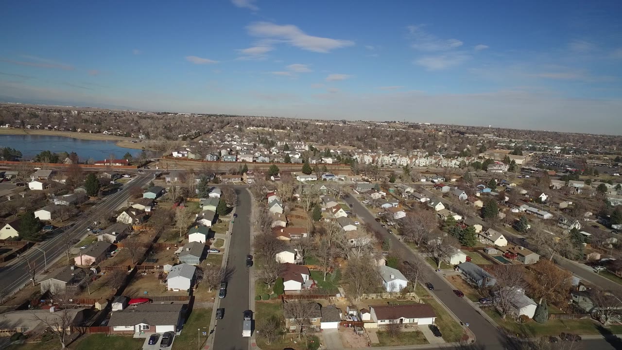 Aerial neighborhood view with houses, roads, and lake under sunny sky.