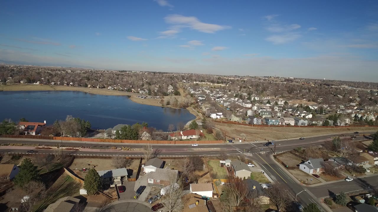 Early aerial frame over Hidden Lake, Colorado, showing the lake left of center, a residential subdivision beyond it, a road intersection below, and no visible people.