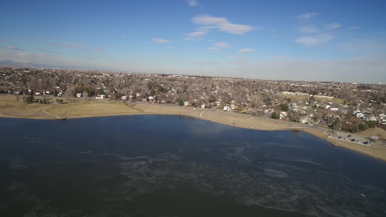 Aerial view over a dark lake toward a shoreline road and residential neighborhood.
