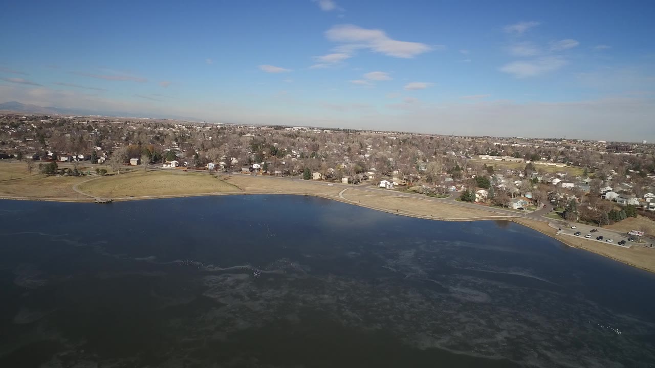 Aerial frame from Hidden Lake, Colorado with the shoreline running across the middle, dark water below, small homes and bare trees beyond, a high horizon, and no visible people.