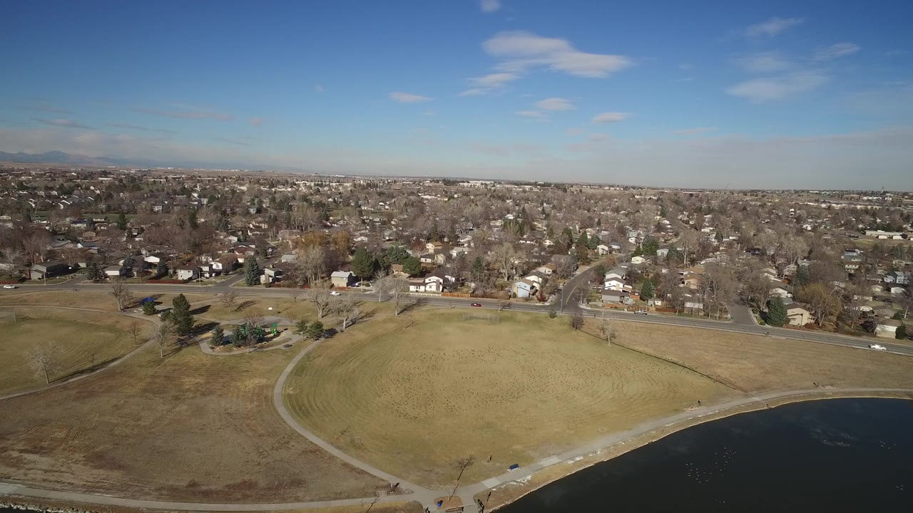 Drone view in Hidden Lake, Colorado showing a broad grass area beside dark water, a pale curving path, a road, residential blocks, and no visible people.
