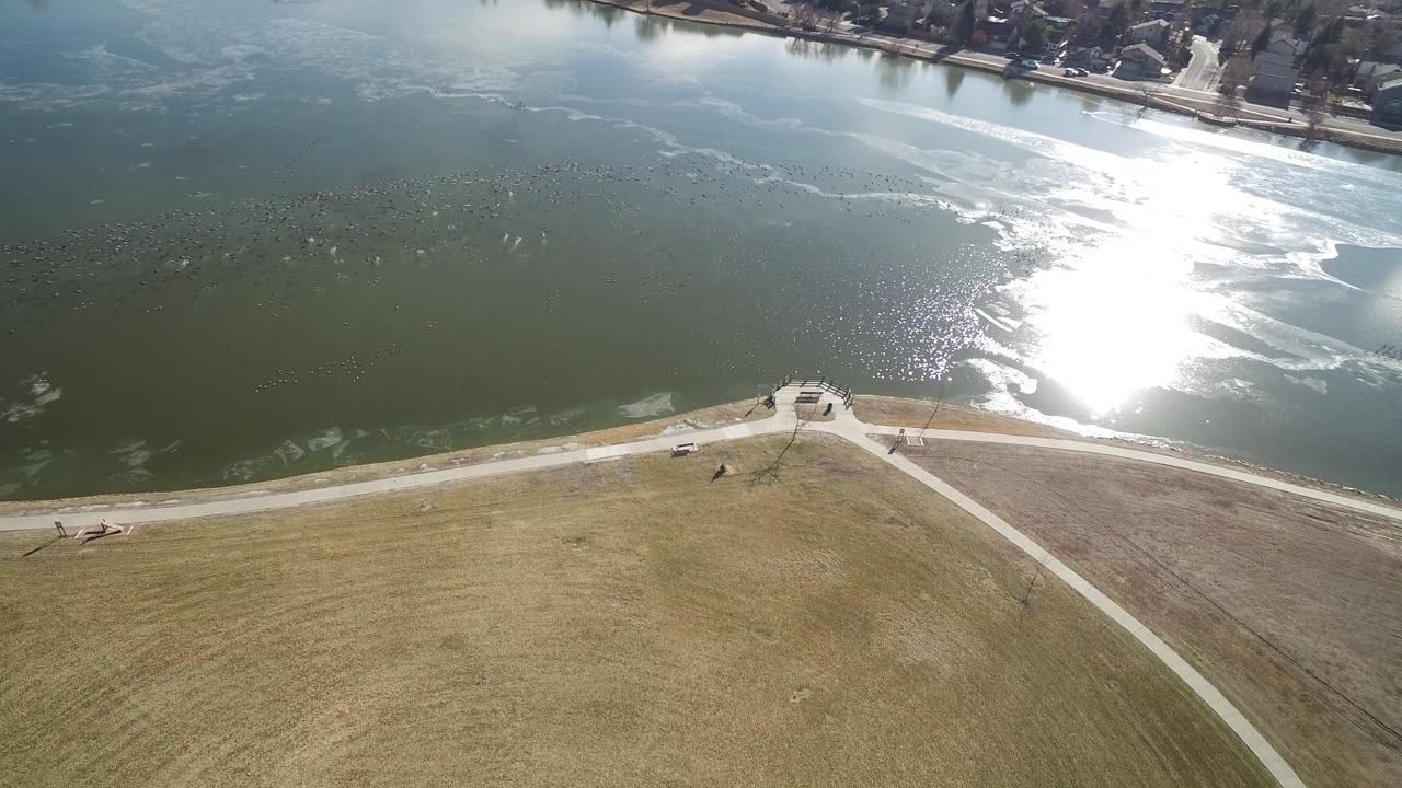 High aerial frame of Hidden Lake Colorado with the shoreline path crossing the lower frame, a small paved lookout near the water, bright reflected sun, and no visible people.