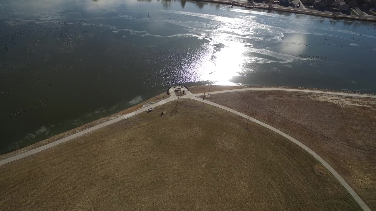 Hidden Lake, Colorado appears from above with the sun reflection shifted across the water, intersecting pale paths below, open grassland, and small human figures near the shoreline.