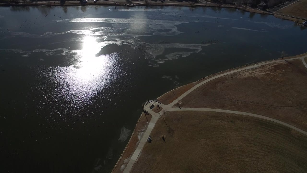 Aerial view of a lake with sun glare, shoreline paths, and brown grass.