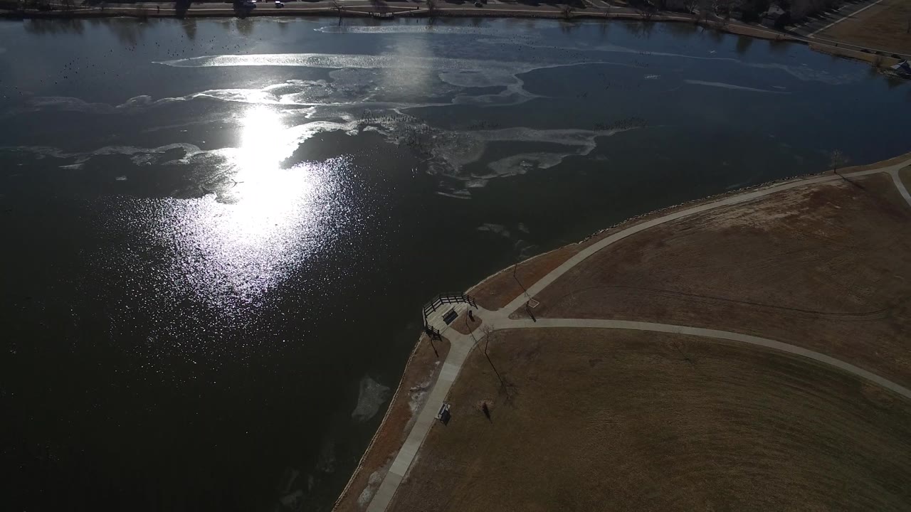 Hidden Lake, Colorado appears from above with bright glare on the water, pale surface streaks, a small shoreline structure, curving paths, grass, and tiny distant figures near the path area.