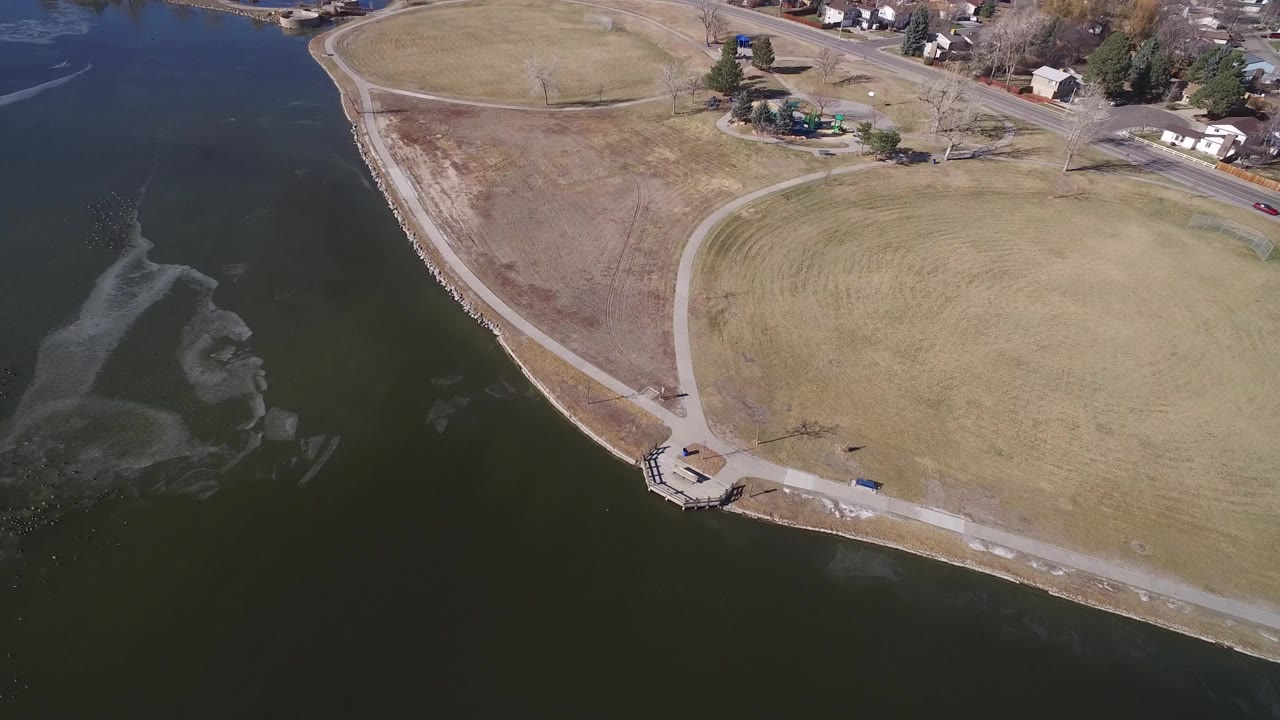 Aerial view over Hidden Lake, Colorado, showing dark water, a curved shoreline, open brown grass, paved paths, a small platform, nearby homes, and no visible people.