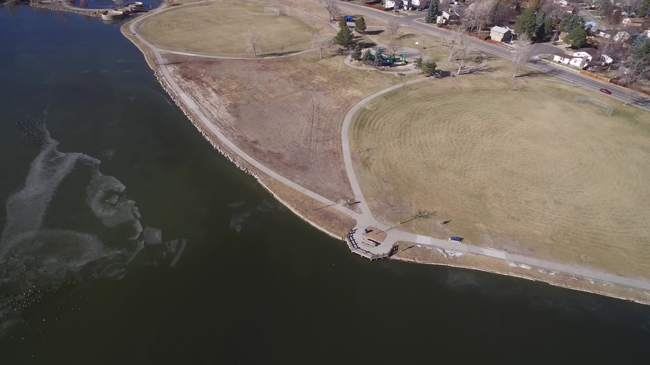 Drone-height view of lake water, shoreline rocks, grass, paths, and a road.