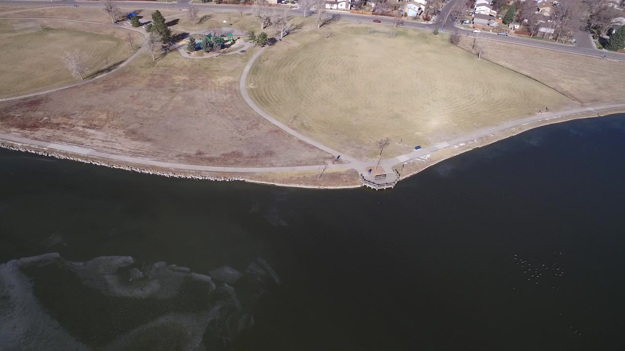 High aerial frame over Hidden Lake, Colorado, showing dark water, a curved stone shoreline, pale grass fields, paved paths, roads, and distant homes with no visible people.