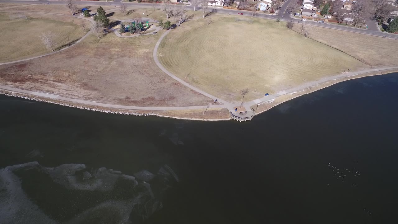 Aerial frame over Hidden Lake, Colorado, with the shoreline running across the center, dark water below, open grass and footpaths above, and residential buildings near the road with no visible people.