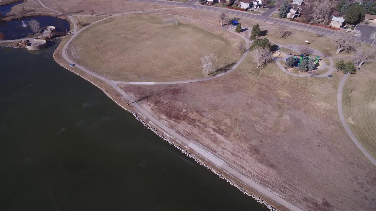 Drone view over Hidden Lake, Colorado, showing dark water along a rocky shoreline, a broad brown field, curving paths, nearby road, houses, and no visible people.
