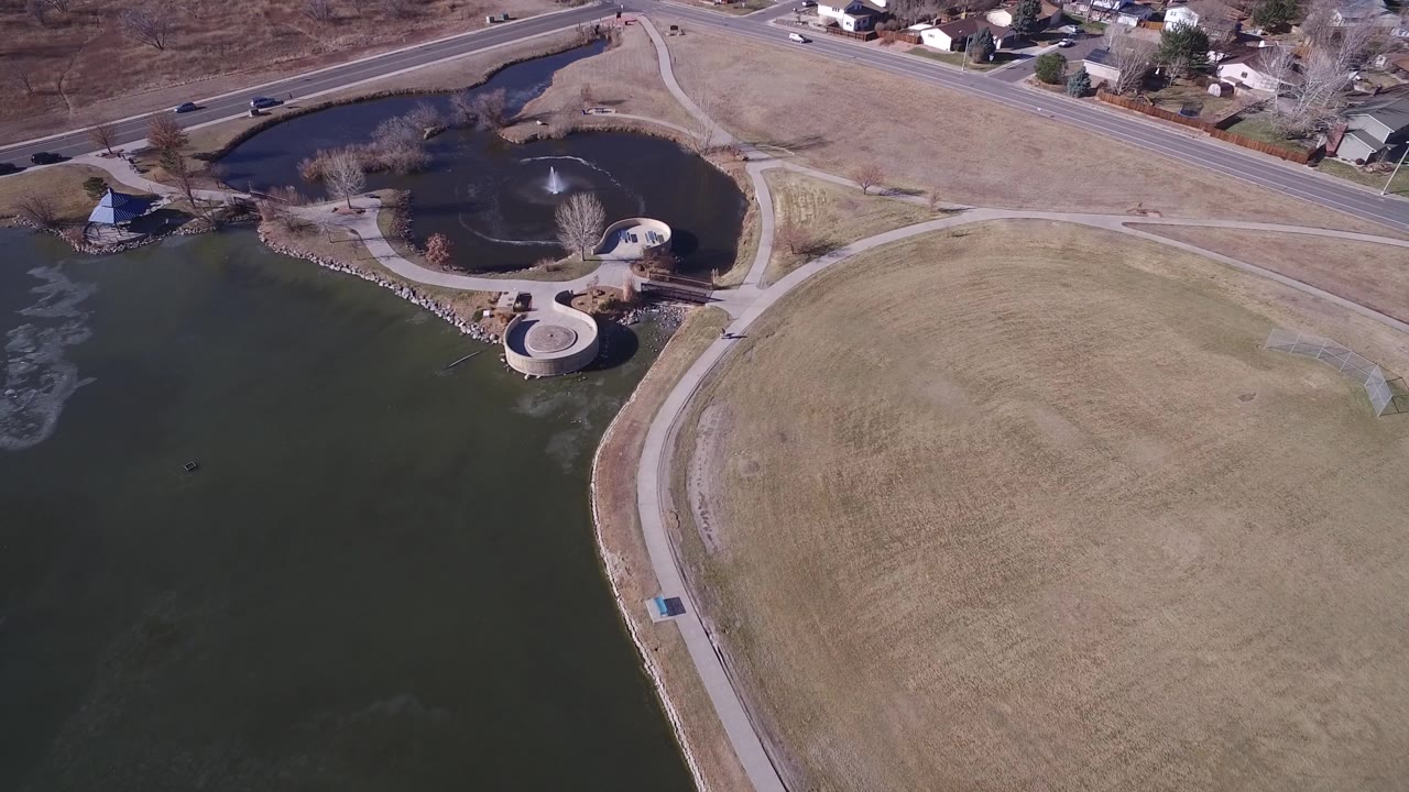 Aerial frame over Hidden Lake, Colorado, with the larger lake filling the lower left, the round pond and fountain near the upper center, dry grass to the right, and no visible people.