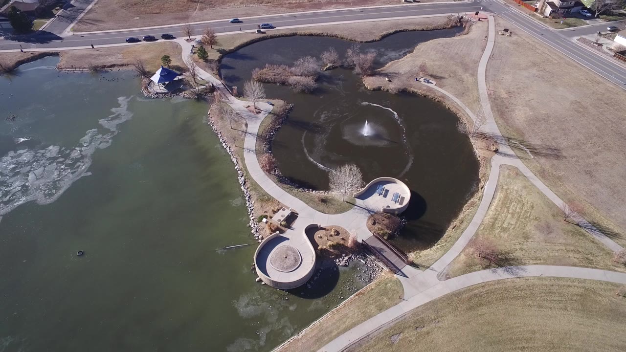 Aerial view of a lake, pond fountain, curved paths, and roadside vehicles.