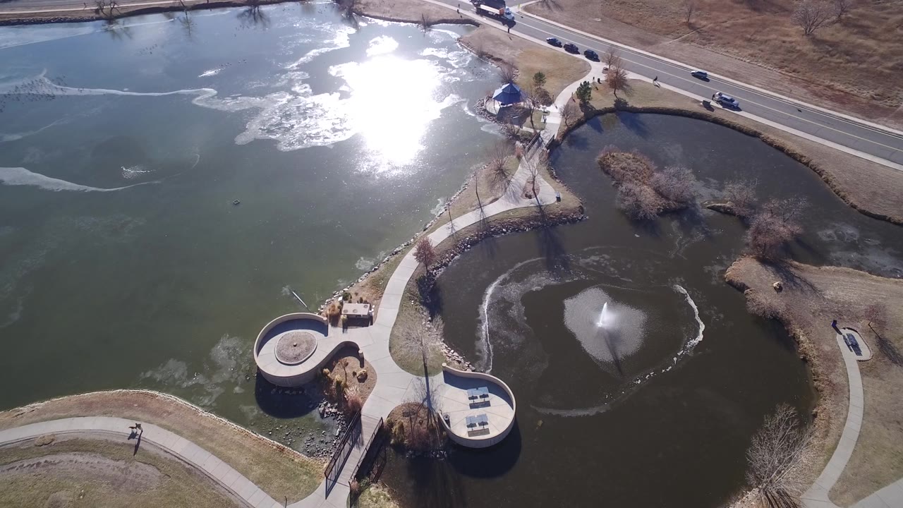 Aerial frame over Hidden Lake, Colorado, with the bright water reflection near the upper center, the fountain pond lower right, circular concrete overlooks at the shore, and no visible people.