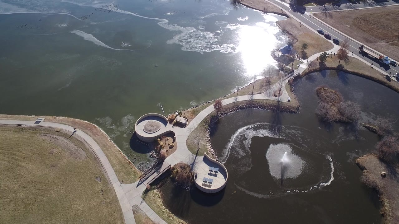 Aerial frame over Hidden Lake Colorado showing green lake water with sun glare, a darker pond fountain, curved paths, a small bridge, circular overlooks, and no visible people.