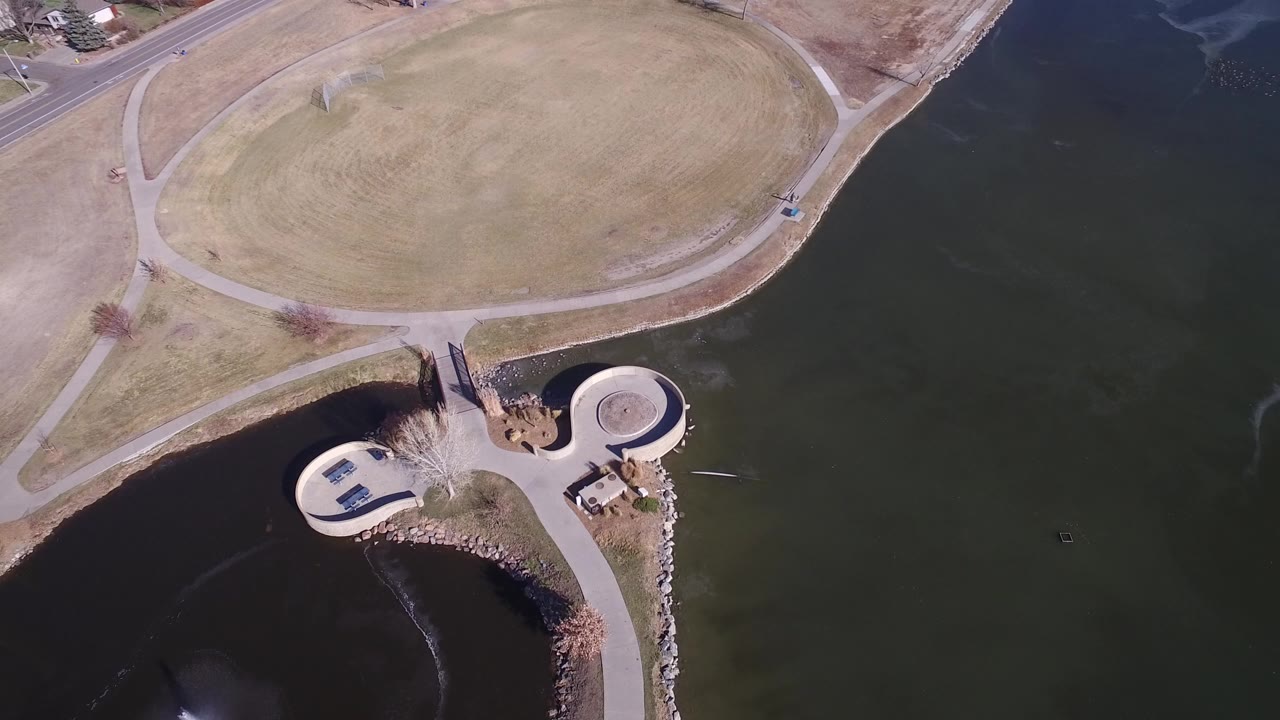 Top-down aerial view of lake water, grass field, paths, and circular shoreline structures.