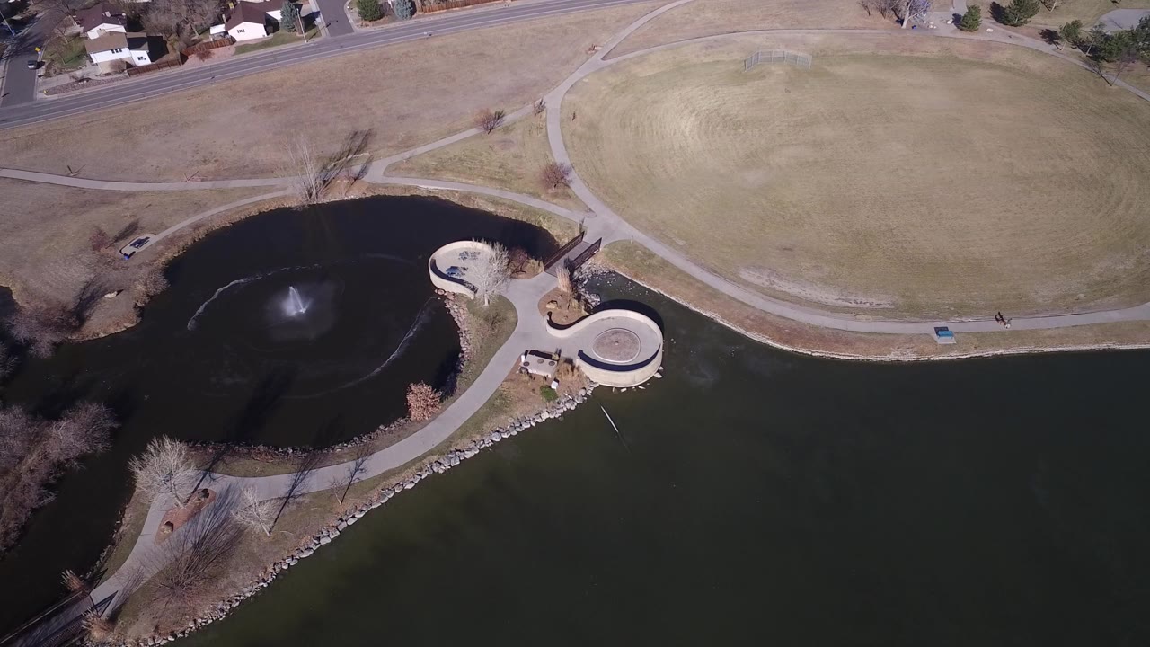 Overhead view of lake water, dry grass, paths, fountain, road, and houses.
