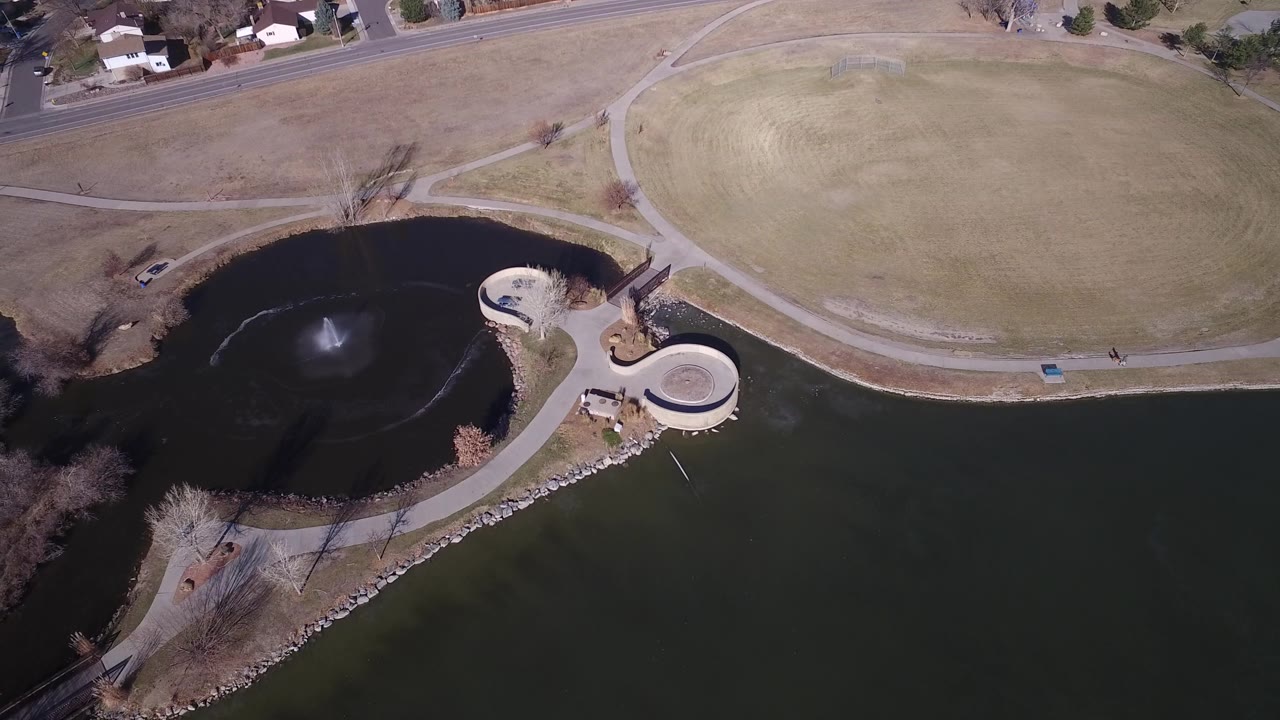 Second overhead frame of Hidden Lake Colorado with the circular concrete platform clearer at right, fountain spray in the pond, dry grassland above, and no visible people.