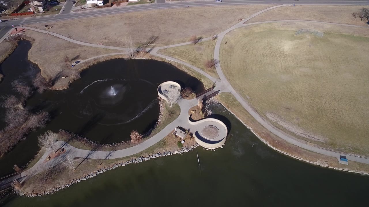 Overhead view of a park pond with fountain, curved paths, circular overlooks, road, and lawns.