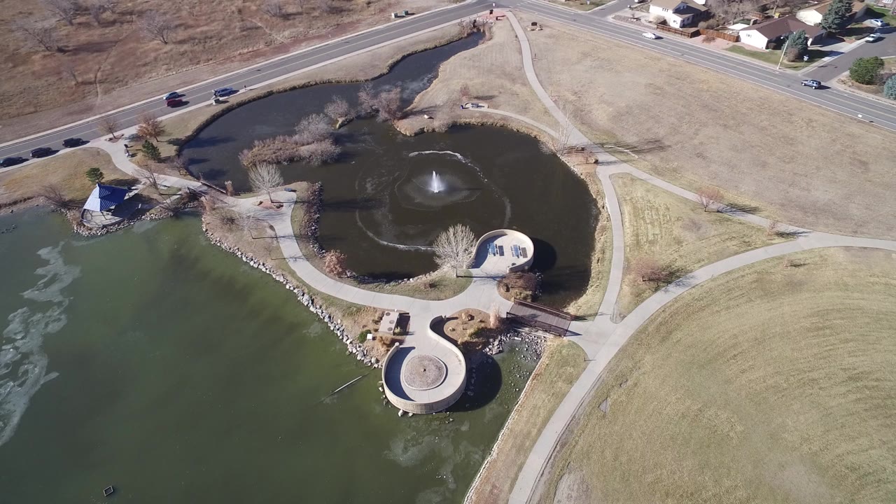 Aerial view of a lake, pond fountain, paths, road traffic, and winter-brown grass.