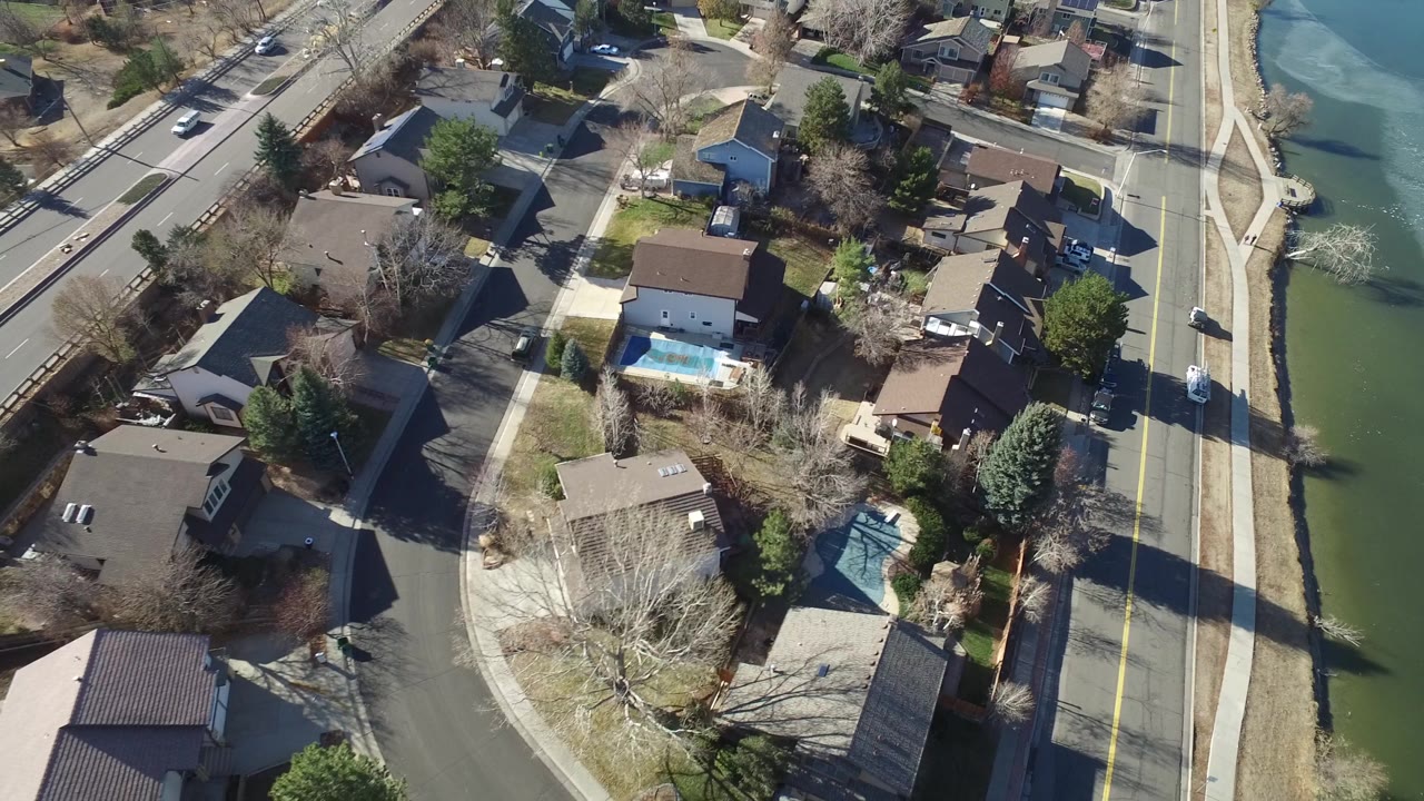Drone-height view of Hidden Lake houses, fenced yards, a bright blue backyard pool, side streets, and the road corridor at frame right, with no visible people.