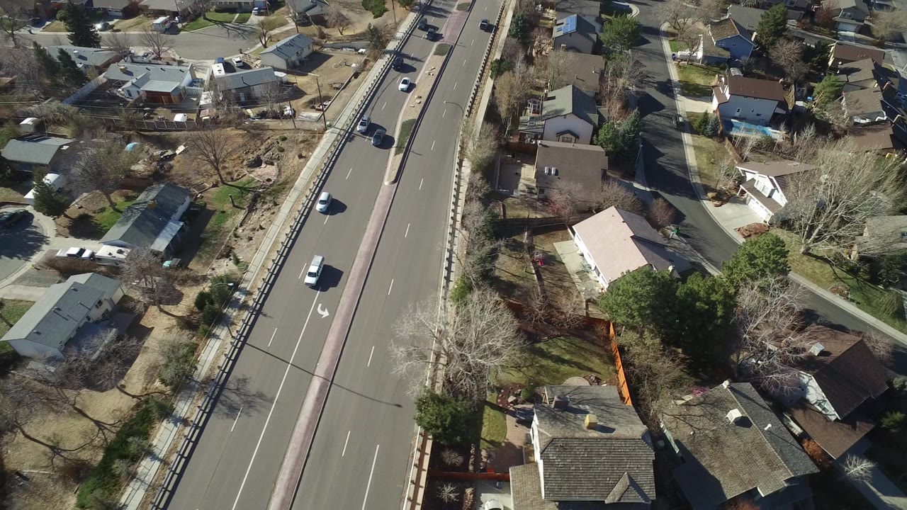 Aerial view in Hidden Lake, Colorado with the divided road closer to center, cars traveling in both directions, and houses and yards spreading along the roadside, with no recognizable people visible.