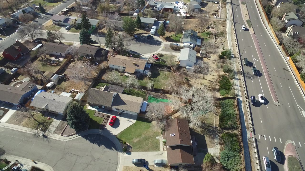 Aerial frame over Hidden Lake, Colorado, with houses and backyards filling the left side while the busy road and several vehicles run up the right edge.