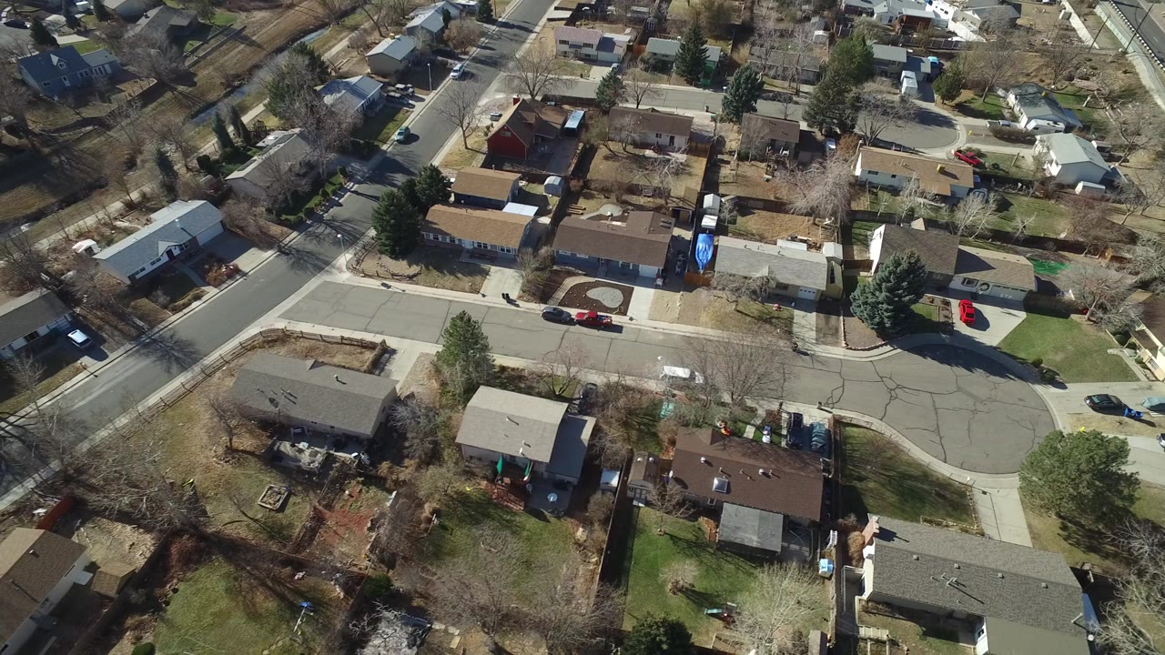 Aerial frame in Hidden Lake, Colorado showing detached homes, small lawns, driveways, backyard fencing, road curves, and parked vehicles with no visible people.