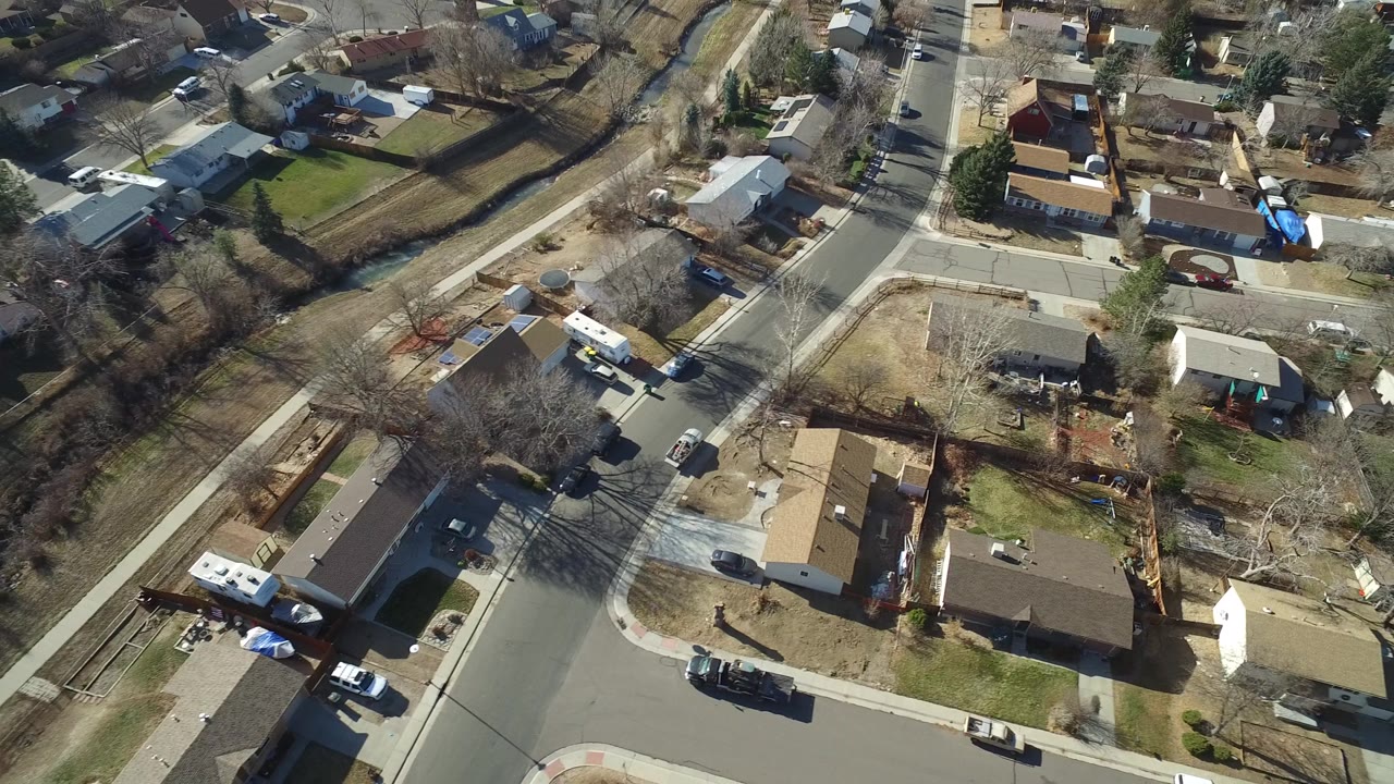 High-angle frame over Hidden Lake, Colorado showing intersecting neighborhood streets, rooftops, driveways, stopped cars, dry trees, and no visible people.