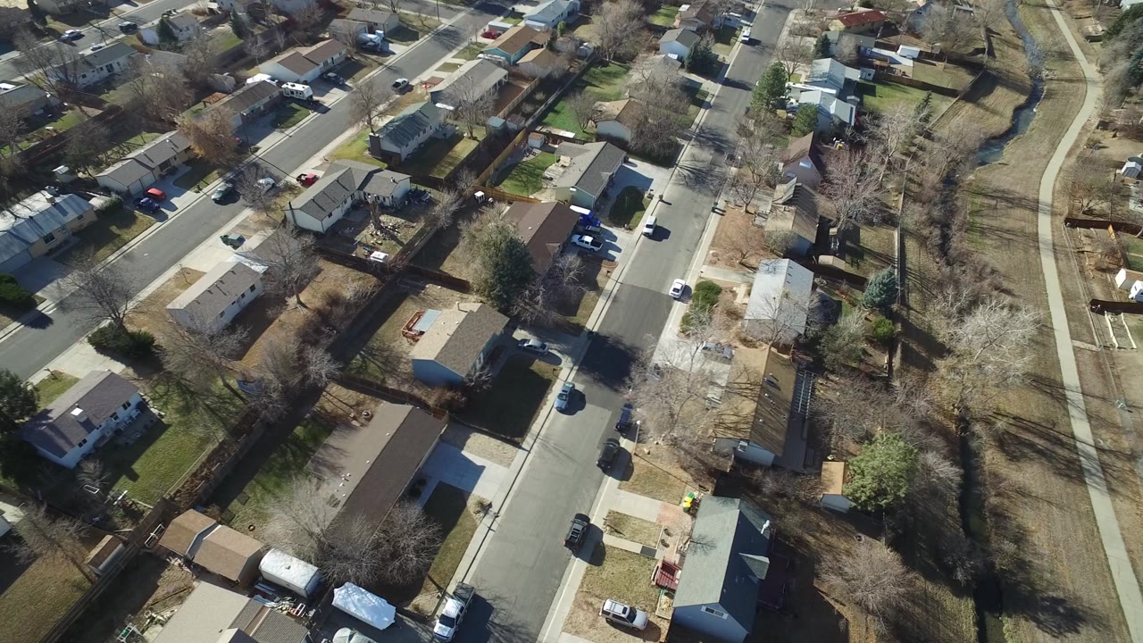 Aerial view of a residential street with homes, trees, driveways, and parked vehicles.