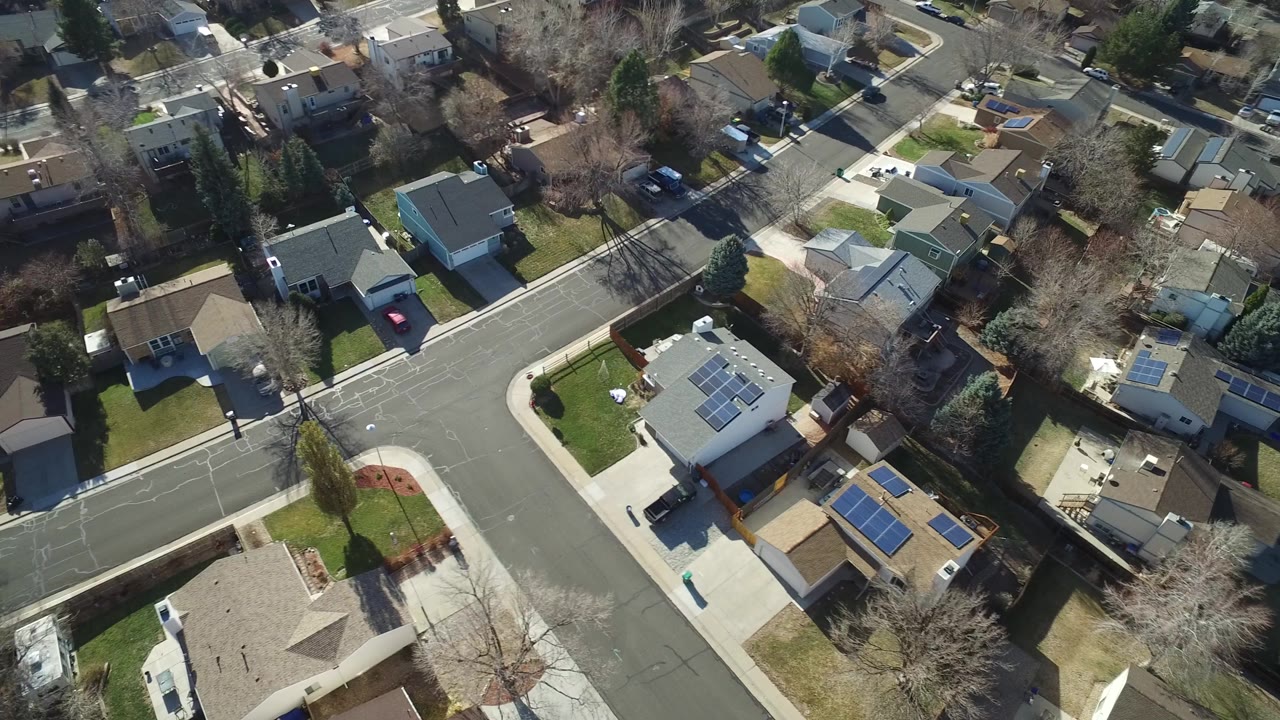 Aerial overhead frame in Hidden Lake, Colorado showing a curved neighborhood road, detached houses, fenced yards, parked cars, solar panels, and no visible people.