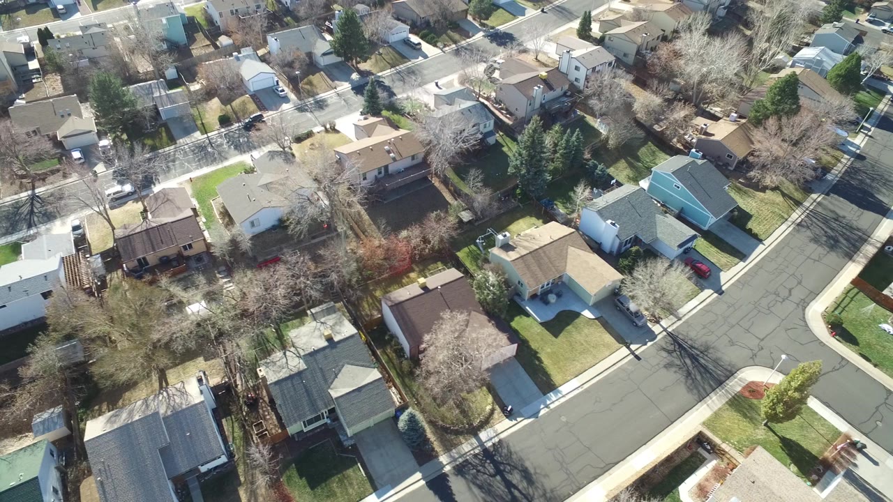Elevated drone frame over Hidden Lake, Colorado homes beside a broad curved street, with lawns, driveways, rooflines, and winter-bare trees visible and no visible people.