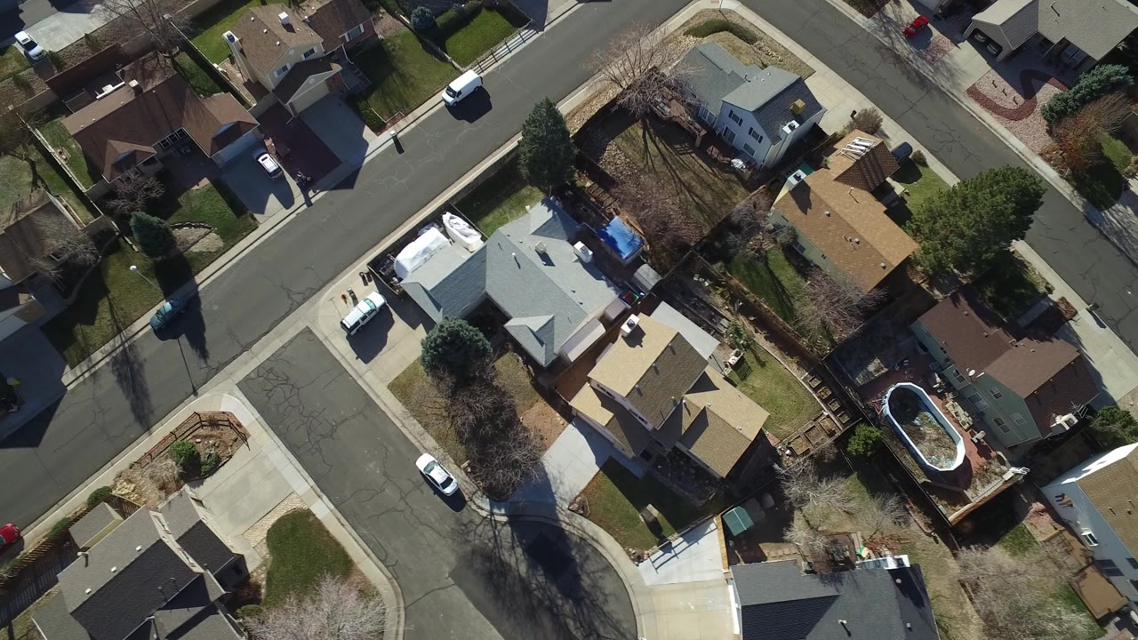 Aerial view of a suburban block with homes, driveways, trees, and vehicles.