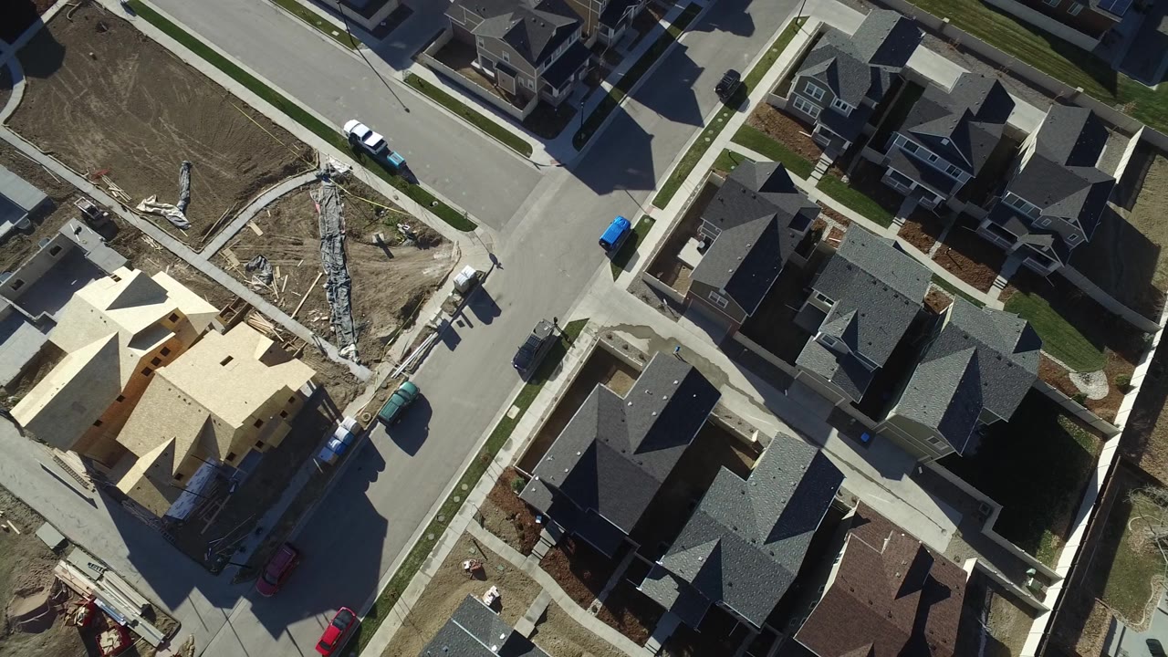 Overhead frame in Hidden Lake, Colorado with the under-construction house shifted lower in view, finished dark-roof homes around it, a road junction, parked cars, dirt ground, and no visible people.