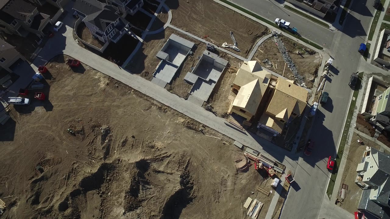 High drone frame over Hidden Lake, Colorado showing a large graded dirt area in the foreground, homes along the upper street, construction framing to the right, and no visible people.