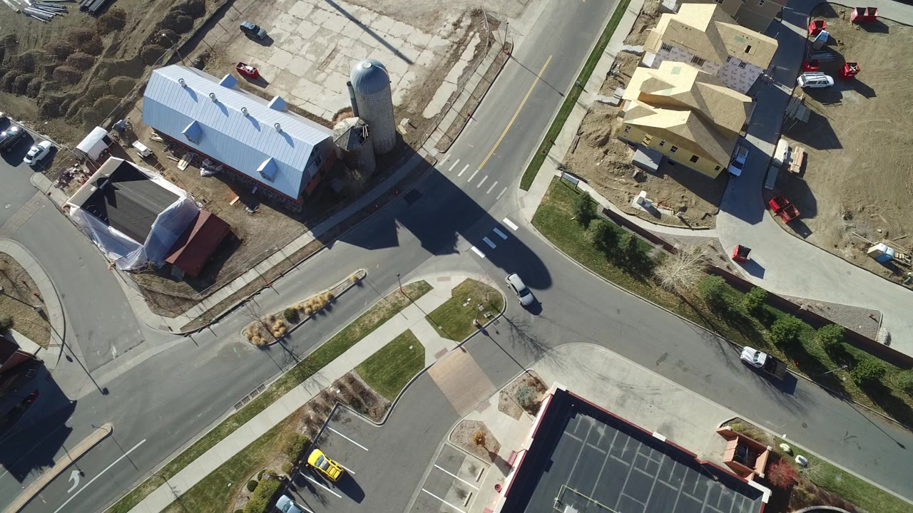 Aerial view of a road intersection with buildings, parked cars, sidewalks, and a tall silo-like structure.