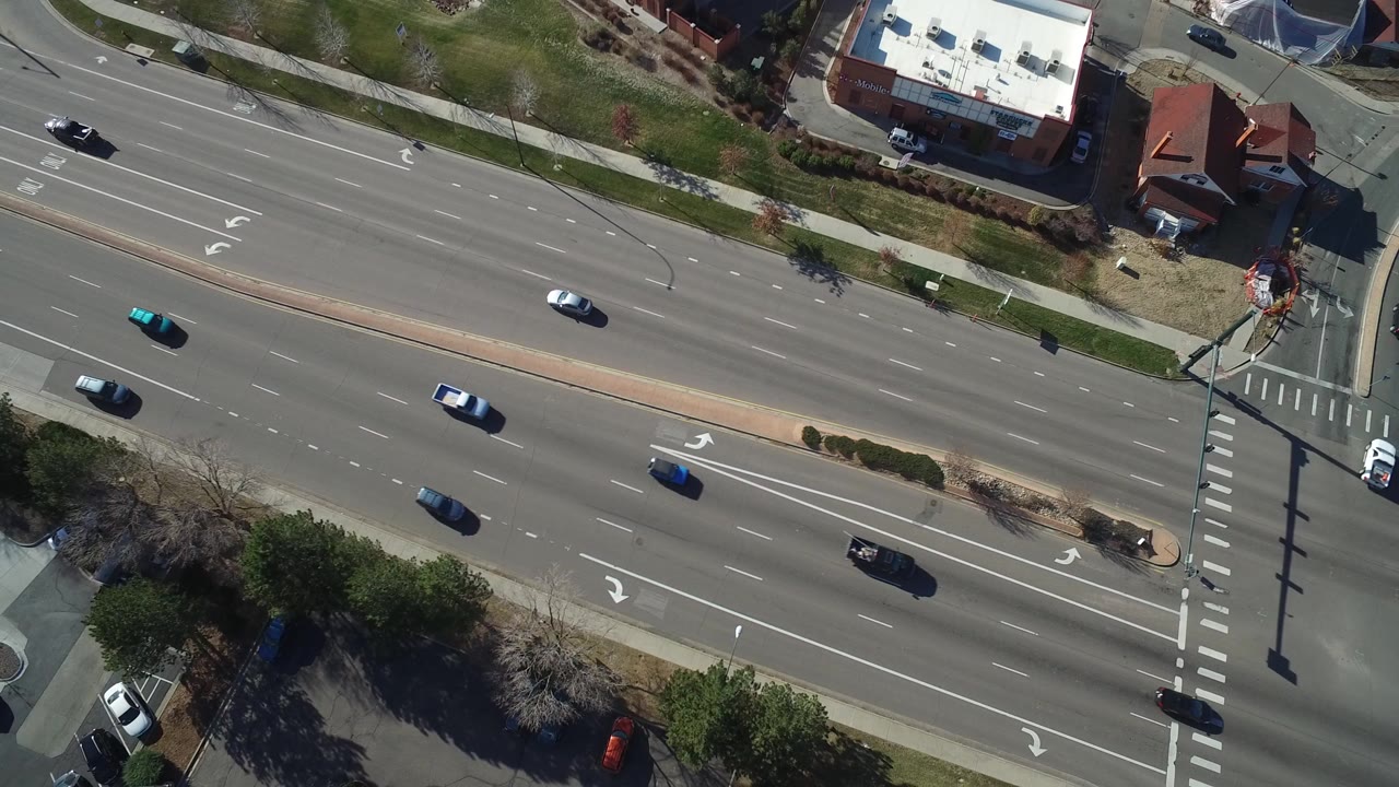 Overhead view of multi-lane road traffic beside parking lots and low buildings.