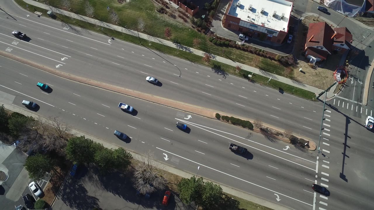 Second overhead frame in Hidden Lake, Colorado, showing cars traveling along a divided road, a landscaped median, sidewalks, parking lot pavement, rooflines, and no visible people.
